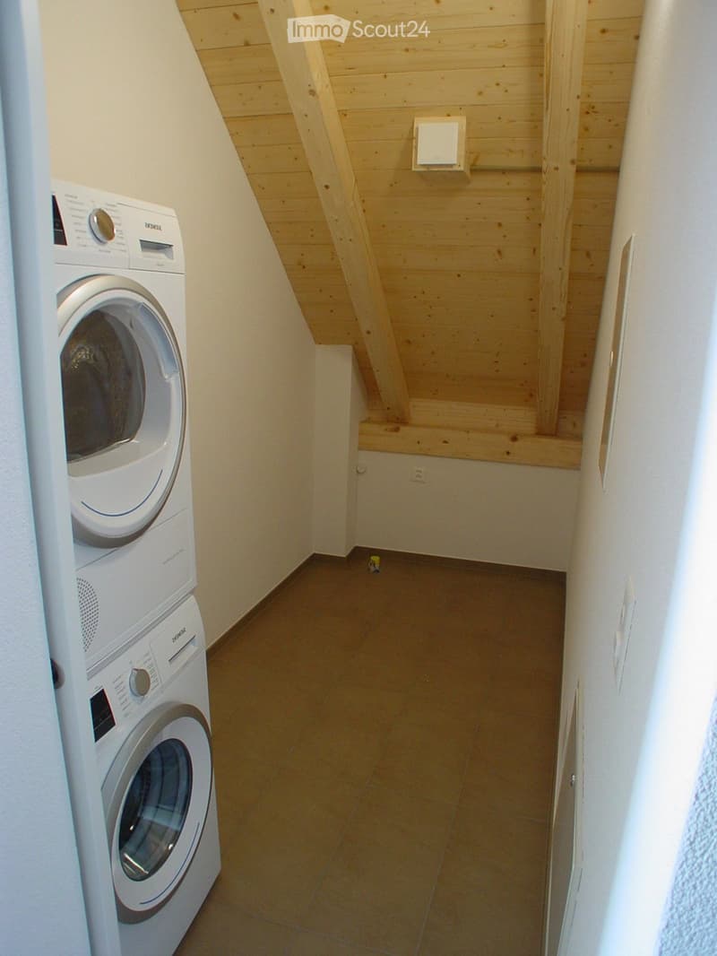 A narrow hallway with wooden slanted ceiling and tiled floor, featuring a stacked washing machine on the left side.
