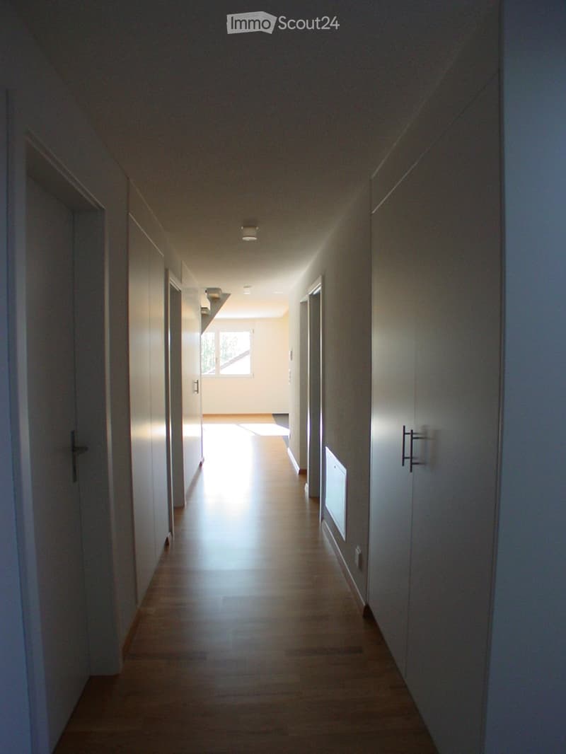 A long, narrow hallway with light-colored wooden flooring and white walls. The hallway features several doors on both sides, with a small window at the far end providing natural light.