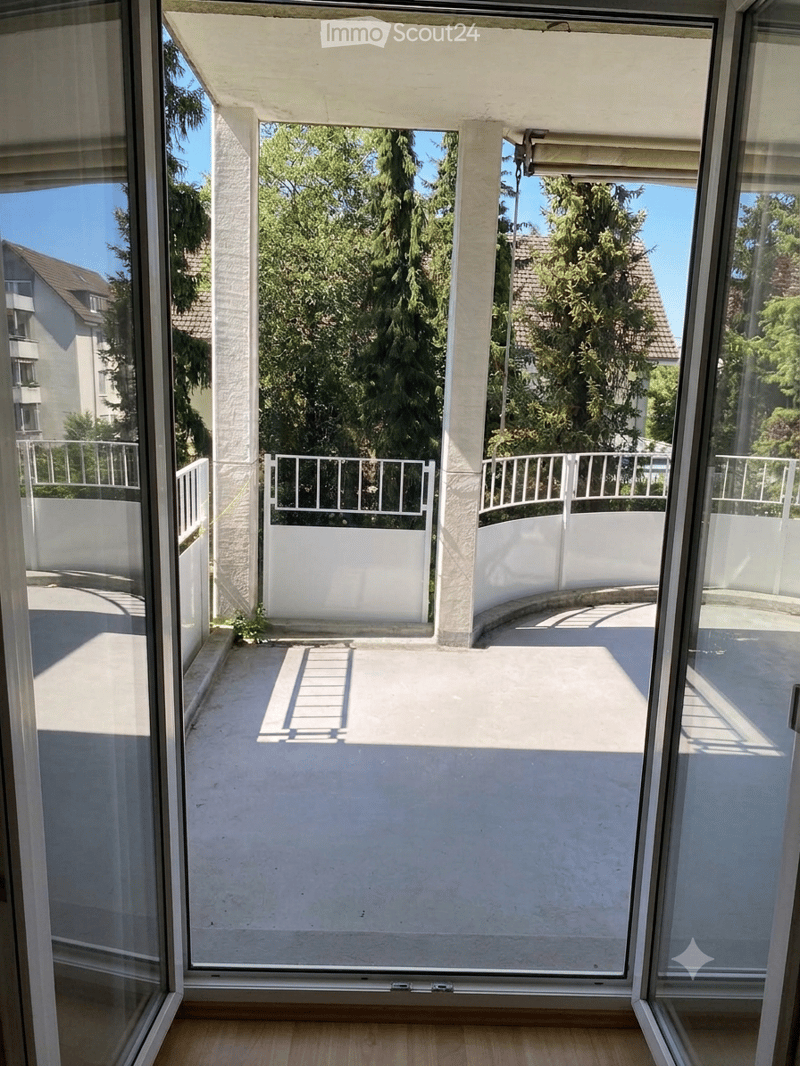 Spacious balcony with white railings, concrete floor, and views of greenery and neighboring buildings.