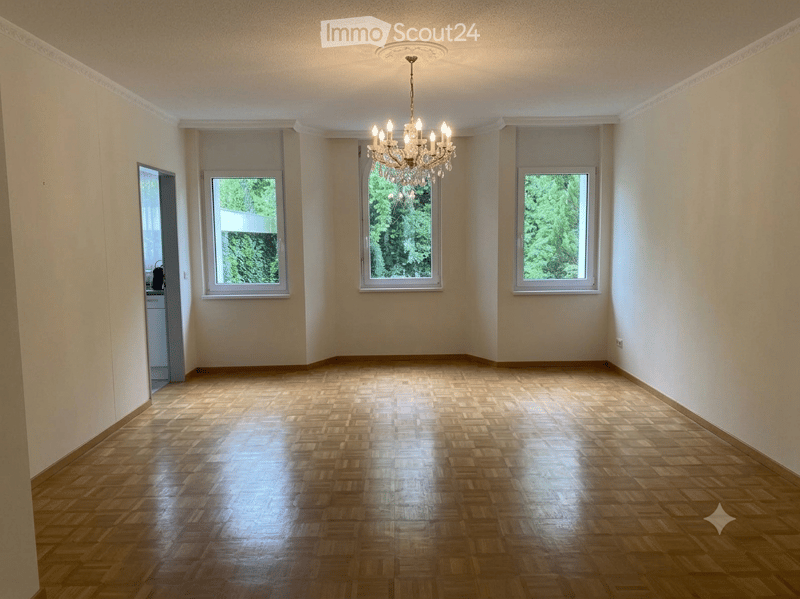Empty room with wooden parquet flooring, white walls, three windows, and a chandelier hanging from the ceiling.
