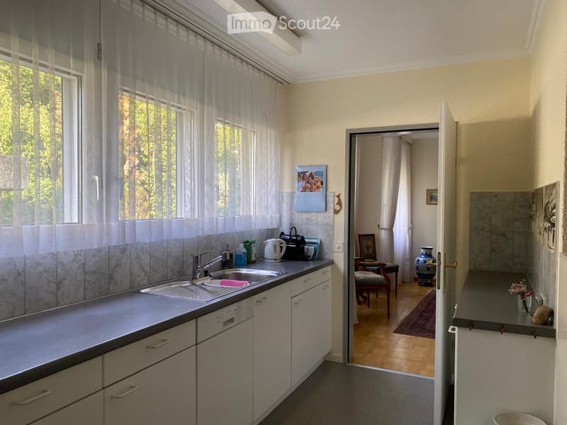 The image shows a modern kitchen with light-colored cabinets, a stainless steel sink, and a dishwasher. A calendar is mounted on the wall, and a small table with a chair is visible in the adjacent room. A vase is placed on the floor near the doorway.