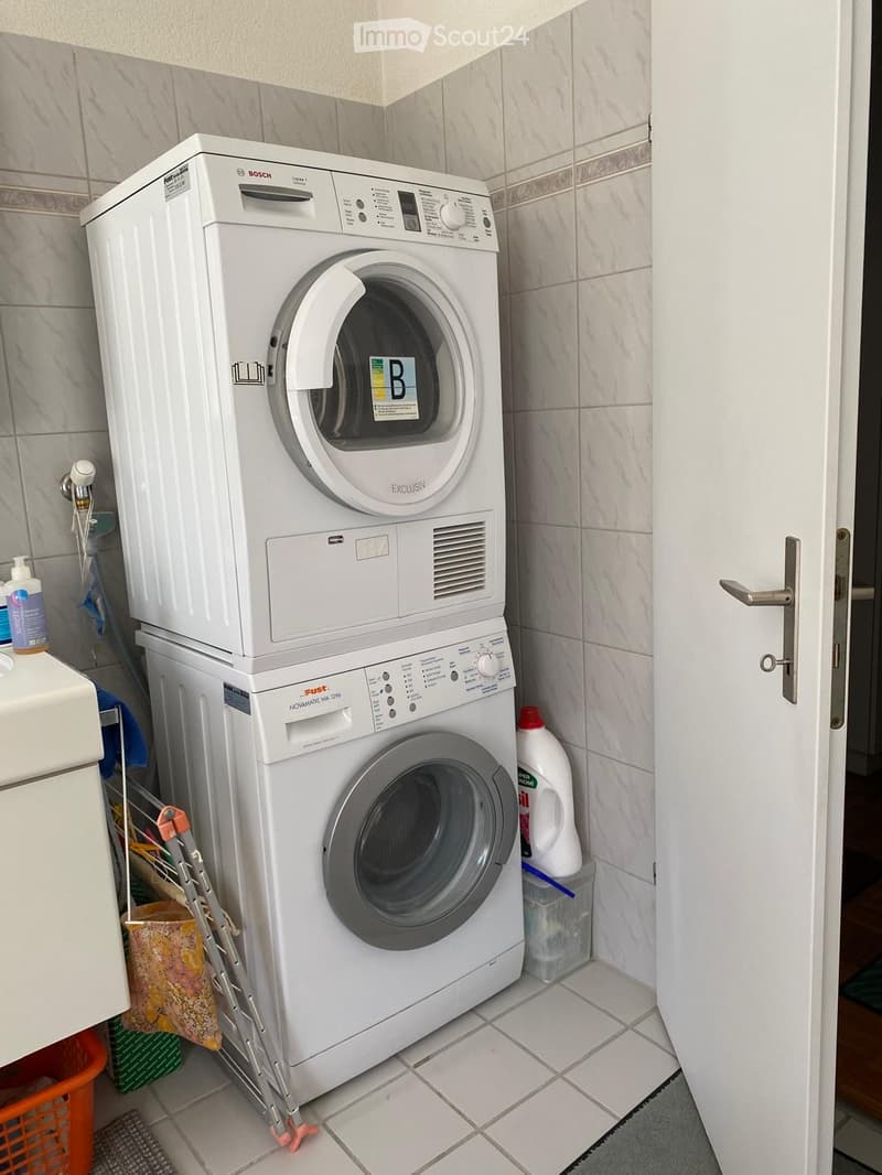 A compact bathroom with a stacked washing machine and dryer, a bottle of detergent, and a laundry basket.