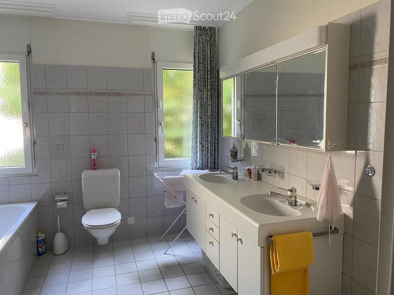 Modern bathroom with a double sink vanity, toilet, bathtub, large mirror, and tiled walls. Includes a window with a curtain and a towel rack with towels.