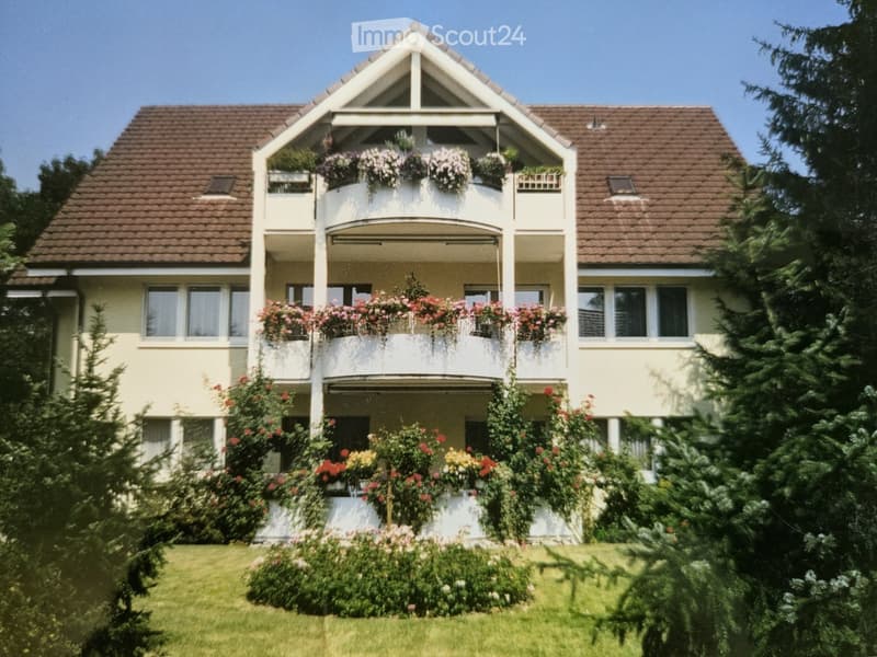 Two-story house with a brown tiled roof, white exterior, multiple windows, and balconies adorned with flower boxes and greenery.