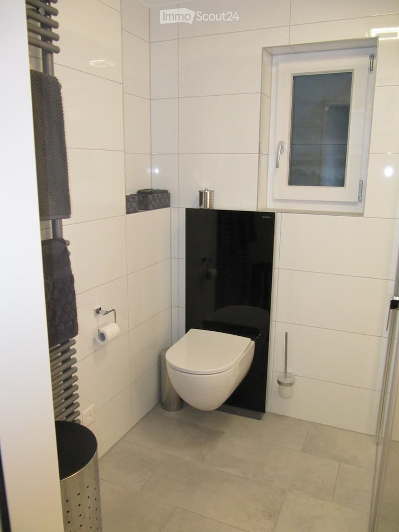Modern bathroom with white tiles, a toilet, a towel rack with towels, a toilet paper holder, a trash can, and a toilet brush holder.