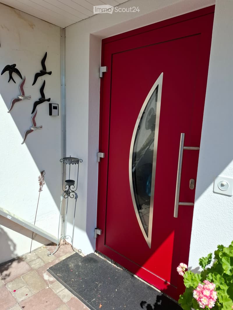 Red front door with a glass panel and silver handle, flanked by white walls and decorative bird sculptures, with a small candle holder and potted plant nearby.