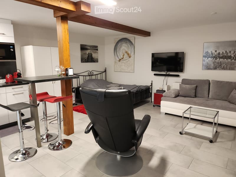 Modern living room with a sofa, coffee table, glass coffee table, black leather chair, bar table with stools, microwave, fridge, and television.