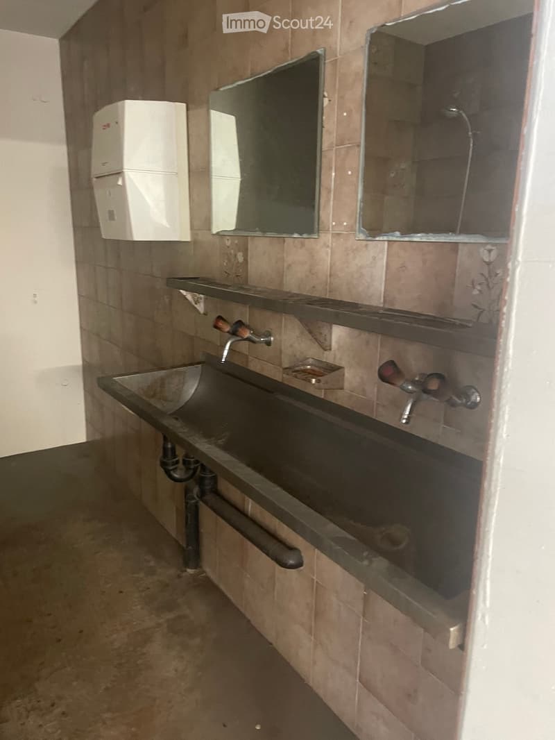 A simple bathroom with a sink, faucet, mirrors, and a toilet paper holder. The walls are tiled in a light brown color, and the floor appears to be concrete.
