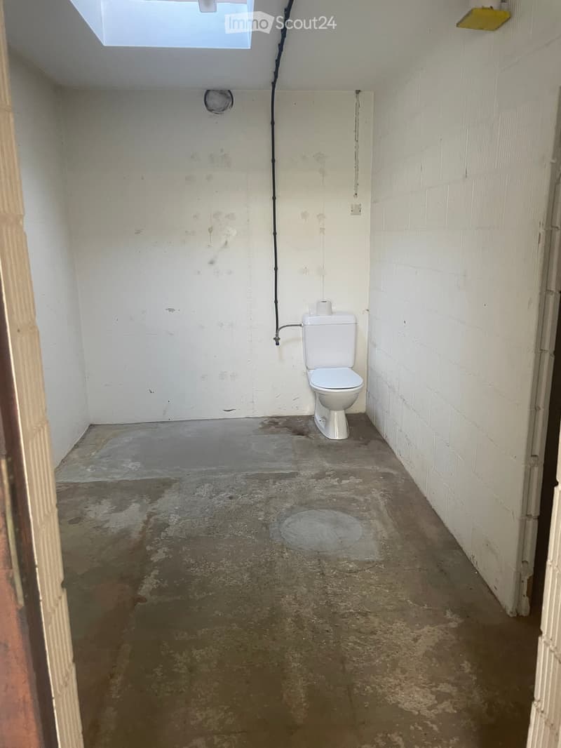Small bathroom with a single toilet, white walls, and a concrete floor showing water damage and stains.