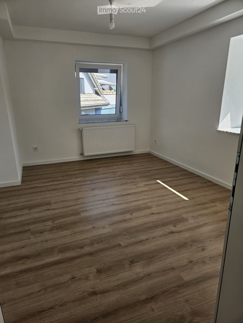 Empty room with wooden flooring, white walls, a radiator, and a window offering a view of a neighboring building.