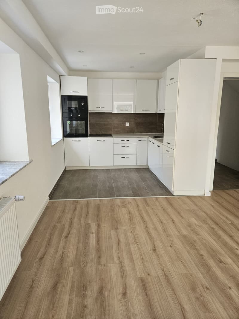 Empty kitchen with white cabinets, dark brown tiles, and wooden flooring. No appliances or furniture are present.