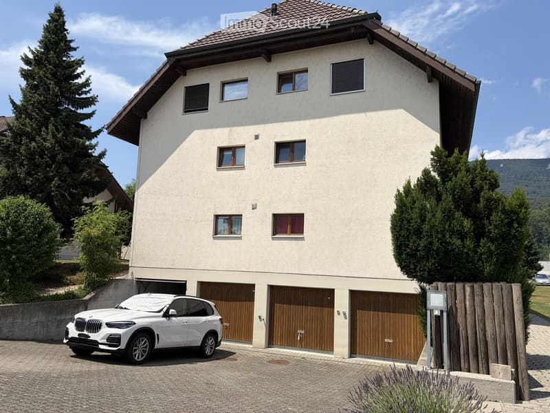 Two-story house with beige siding, multiple windows, and a large garage with wooden doors. A white SUV is parked in front of the garage.