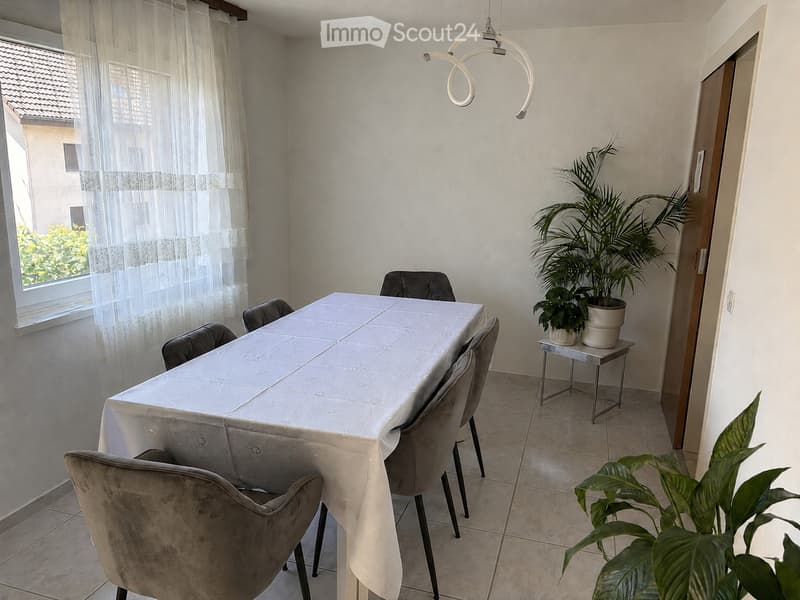 Dining area with a table covered in a white tablecloth, surrounded by four upholstered chairs. A potted plant sits on a small side table near the window, which has sheer curtains. The room has tiled flooring and a modern pendant light fixture hanging from