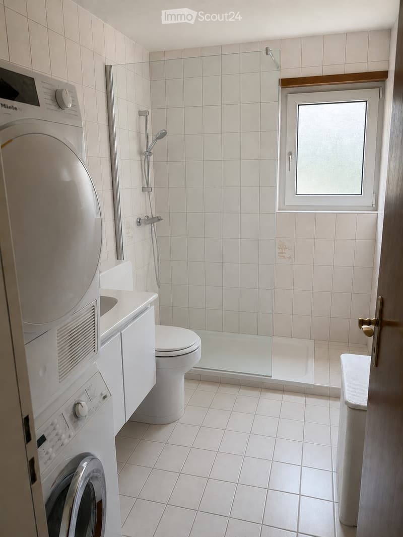 Modern bathroom with white tiles, a toilet, a washing machine, a glass-enclosed shower, and a small window.