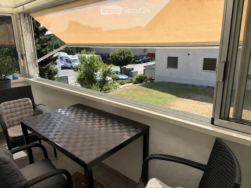 Balcony with a small mosaic-tiled table and two wicker chairs, large windows with a retractable awning, and a view of a parking area with cars and greenery.