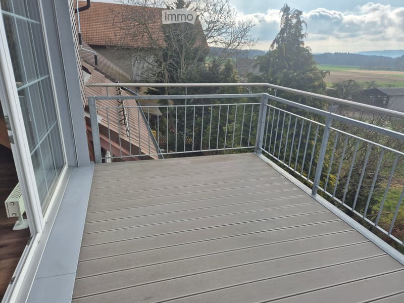 Balcony with wooden decking and metal railing, overlooking a rural landscape with trees and houses.