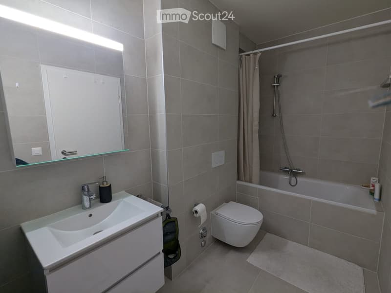 Modern bathroom with white tiles, a white sink, a mirror, a toilet, and a bathtub with a shower curtain. The sink has a faucet and a soap dispenser. There is a towel on the towel rack and a power outlet on the wall.