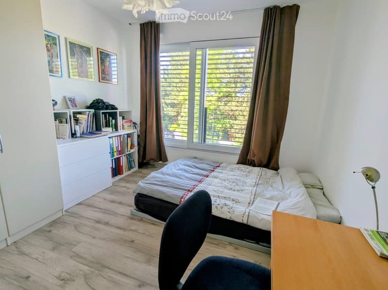 A small furnished room with a bed, desk, chair, and bookshelf. The room has a wooden floor and white walls. The bed is made with a white and red-patterned blanket. The desk has a lamp and a book on it. The bookshelf is filled with books and other items. T