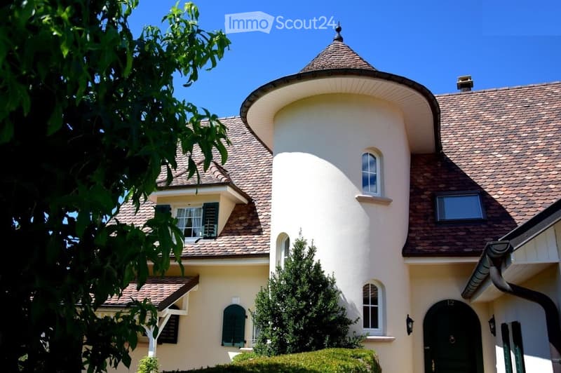 A two-story house with a distinctive round tower, beige exterior, and brown tiled roof. The house features multiple windows with shutters and a well-maintained garden.