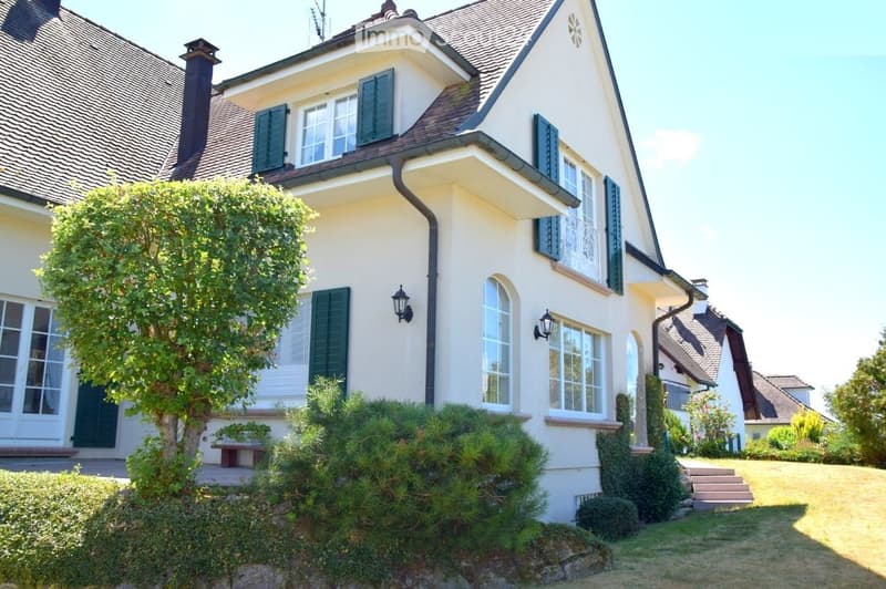 Two-story house with light yellow exterior, dark green shutters, multiple windows, and a well-maintained garden with shrubs and a tree.