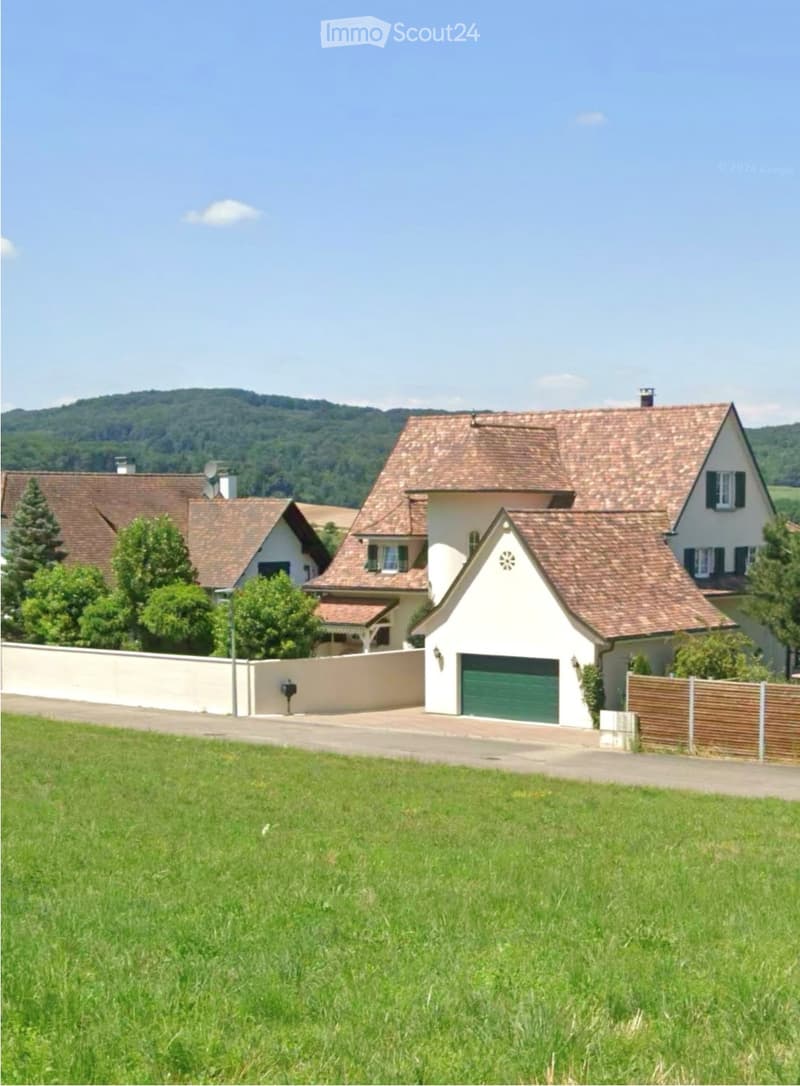 A two-story house with a red-tiled roof, white walls, and a green garage door, surrounded by a well-maintained lawn and trees.