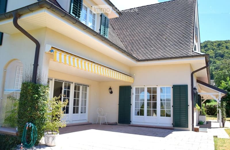 Two-story house with a cream-colored exterior, green shutters, and a yellow striped awning over the entrance. Features include large windows, a white plastic chair on the patio, and a small garden area with a potted plant and a coiled green hose.