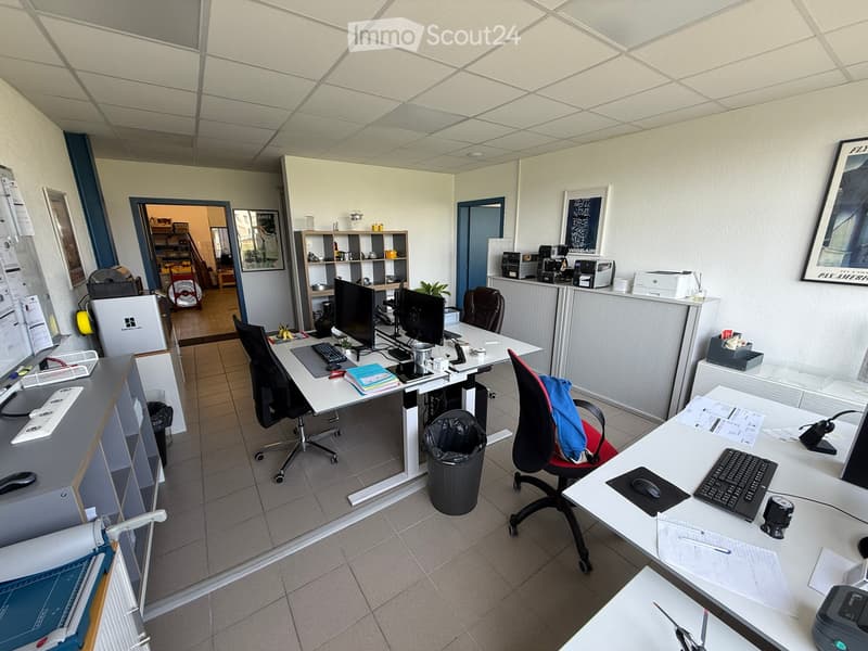 Office space with desks, chairs, computers, printers, and shelves. The room features tiled flooring and white walls.