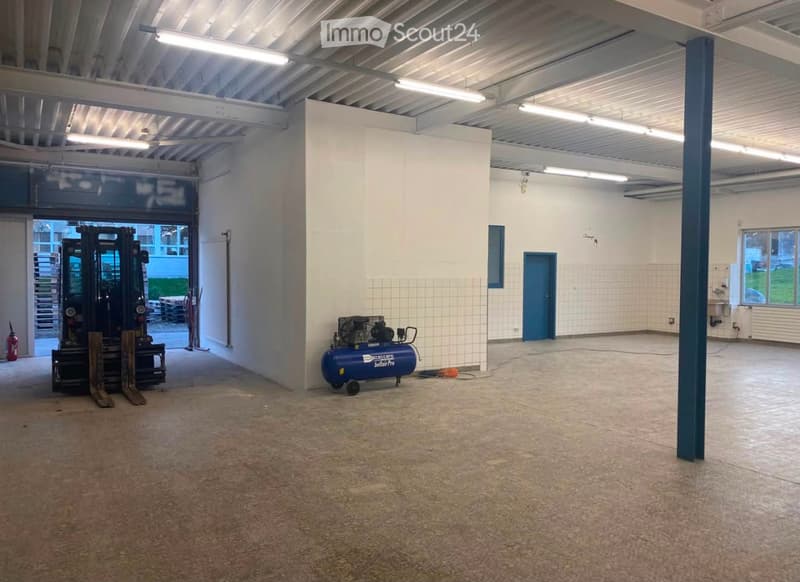 Empty industrial-style room with white walls, concrete floor, and large windows. Contains a forklift, air compressor, and a fire extinguisher.