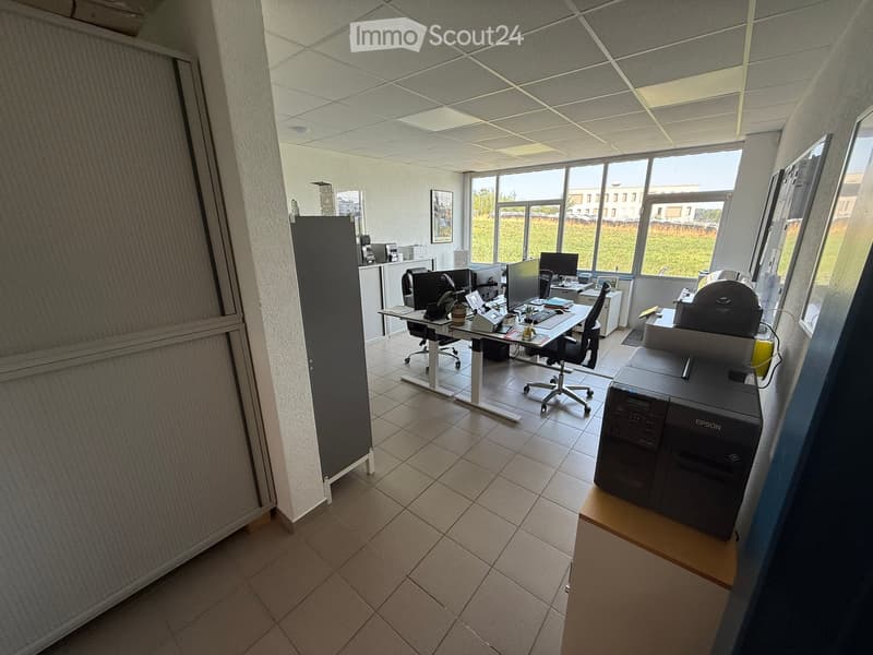 Office space with desks, chairs, computers, printers, and a large window offering a view of the outdoors.
