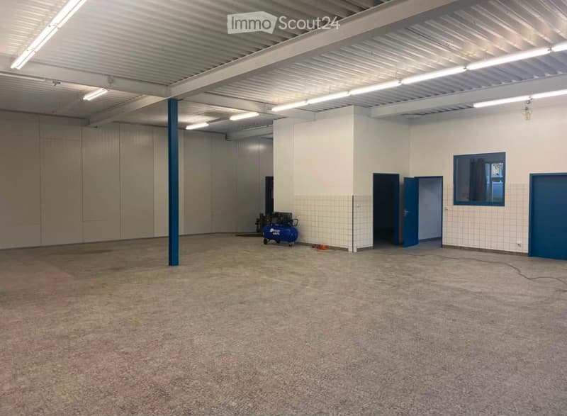 Empty warehouse space with concrete floor, white walls, industrial lighting, and a blue air compressor in the corner.