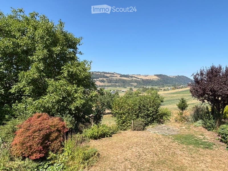 A well-maintained garden with a variety of trees, shrubs, and a clear view of rolling hills in the background.