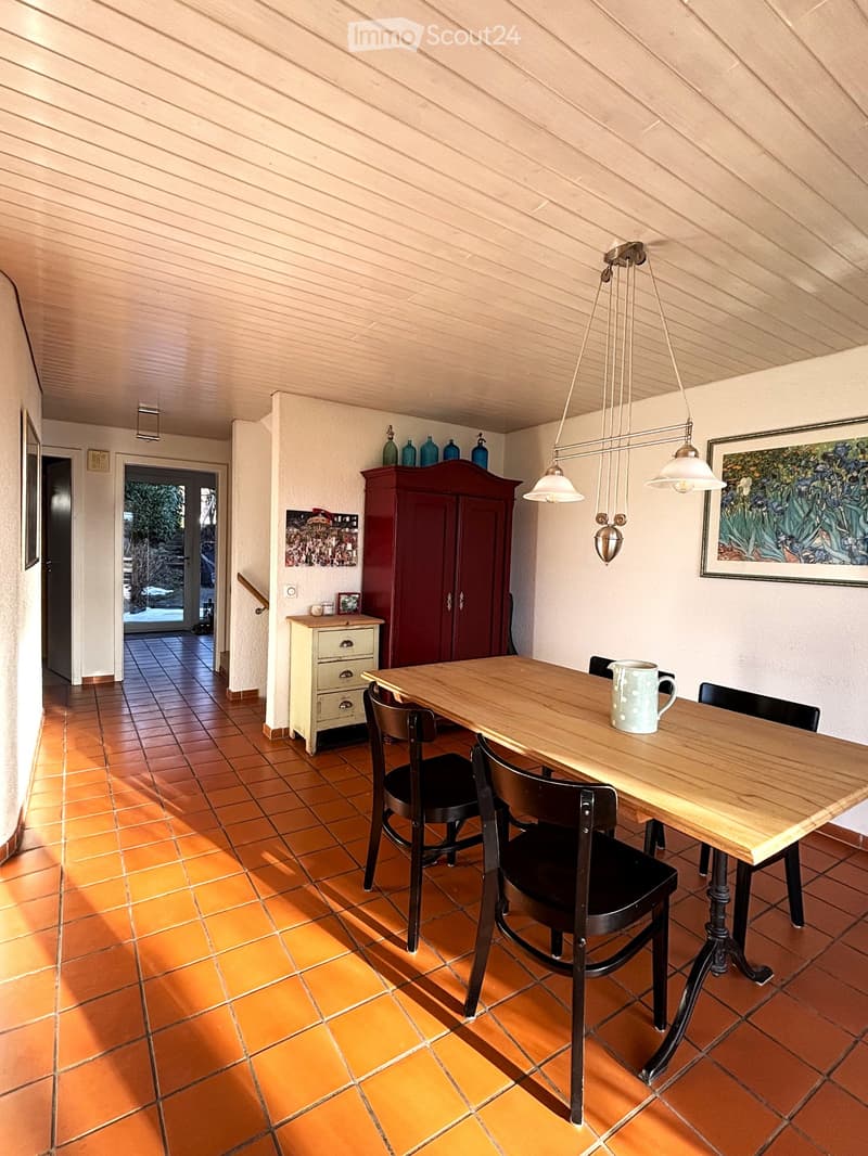Dining room with a large wooden dining table, chairs, wooden floor, chandeliers, side table, red cabinet, cabinet with drawers, painting on the wall, wooden door