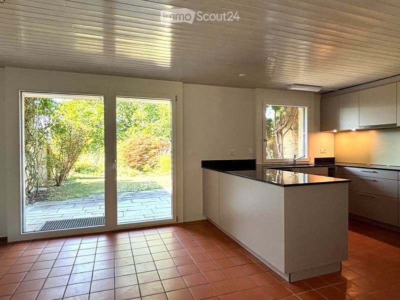 Modern kitchen with a central island, built-in cabinets, and a sink, featuring large windows and sliding doors leading to an outdoor area.