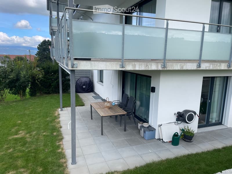 Outdoor patio area with a wooden table, chairs, a basket, potted plant, garden hose, and storage boxes. The patio is tiled and adjacent to a grassy lawn, with a covered barbecue and a modern balcony above.