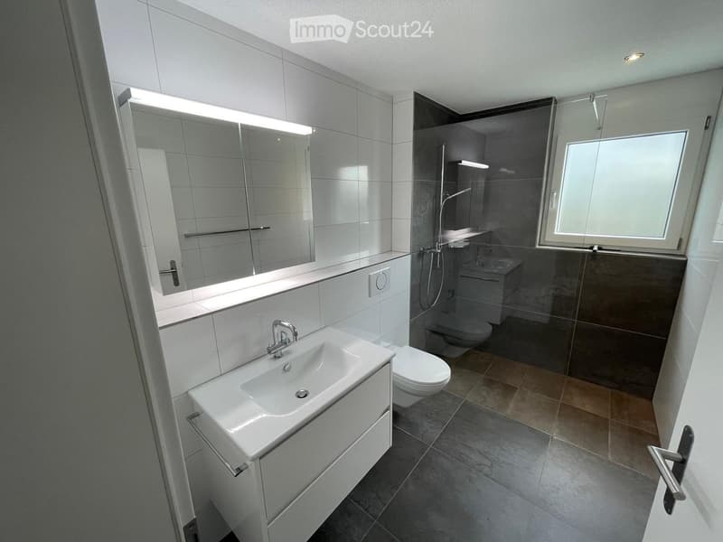 Modern bathroom with white tiles, a sink with a mirror, a toilet, and a glass-enclosed shower area with a window.