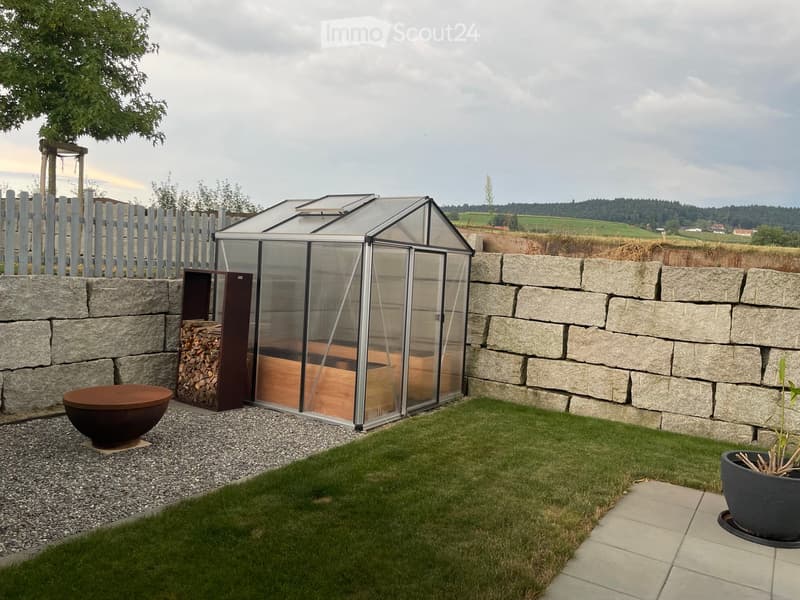 Well-maintained garden with a greenhouse, stone wall, neatly trimmed grass, and decorative pots.