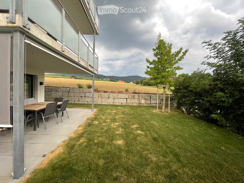 Outdoor patio area with a wooden table and chairs, surrounded by a grassy lawn and a stone wall, with a view of a field and trees in the background.
