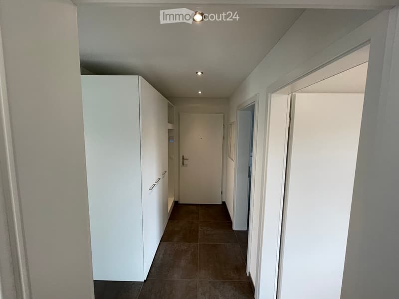 A clean, modern hallway with tiled flooring, white walls, and recessed lighting. Features include built-in white cabinetry on one side and several doors leading to other rooms.