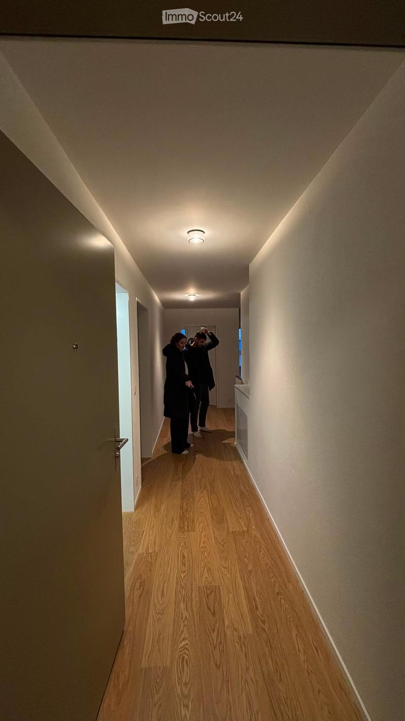 A narrow hallway with wooden flooring, beige walls, and recessed ceiling lighting. Two individuals are walking down the hallway.