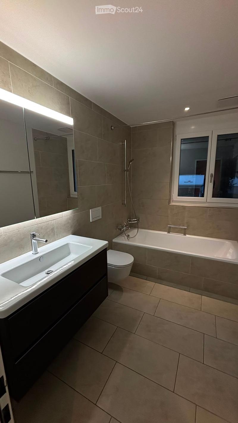 Modern bathroom with beige tiles, a large sink with a chrome faucet, a toilet, a bathtub, and a large mirror with built-in lighting.
