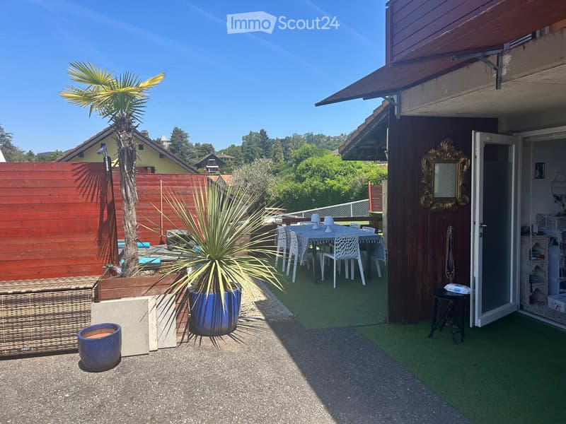 Outdoor patio area with artificial grass, a small table and chairs, potted plants, and a wicker basket. The patio is adjacent to a house with an open sliding door.