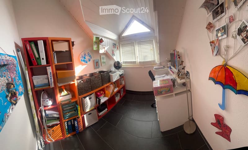 A cozy, well-lit room with a slanted ceiling and skylight. Features a large bookshelf filled with books and storage bins, a desk with a chair, and various decorative items including a colorful umbrella and a globe.