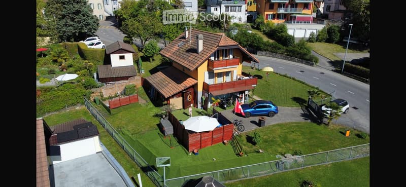 Aerial view of a two-story house with a tiled roof, surrounded by a well-maintained garden, a garage, and several parked cars.