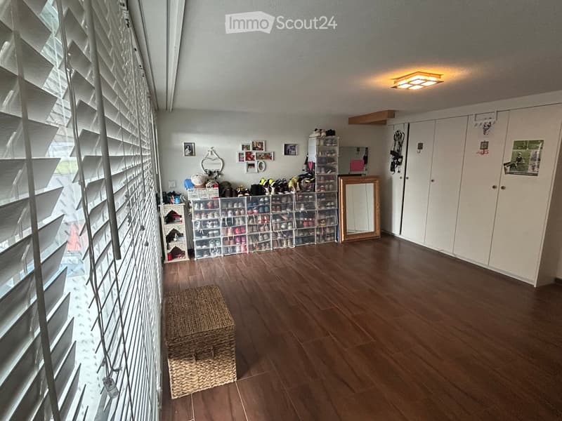 Well-lit hallway with wooden flooring, large window with blinds, shoe rack, mirror, storage cabinet, and picture frames on the wall.