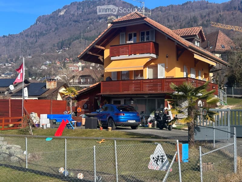 Two-story yellow house with red wooden balconies, surrounded by a fenced yard with a blue car, motorcycle, and various outdoor items.