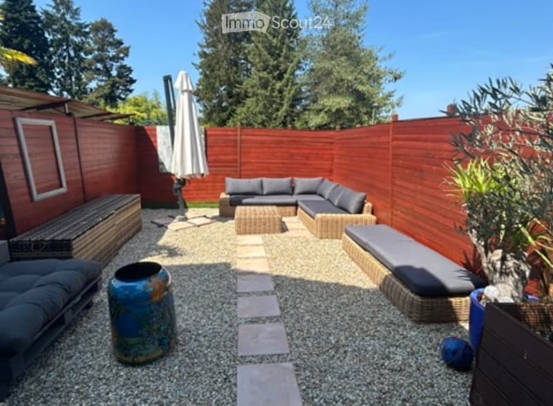 A well-maintained garden with a seating area featuring a sofa, table, and umbrella, surrounded by wooden fencing and lush greenery.