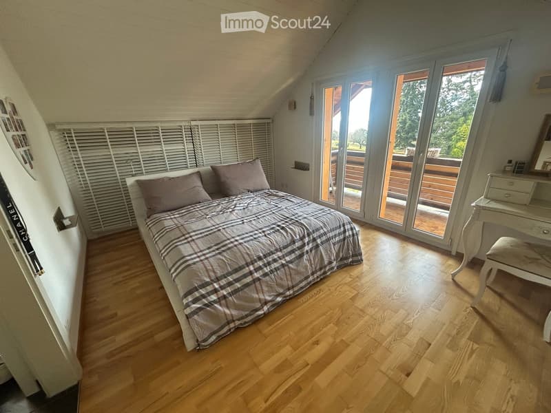 Bright bedroom with wooden flooring, a large bed with plaid bedding, a small desk with a stool, and large windows opening to a balcony.