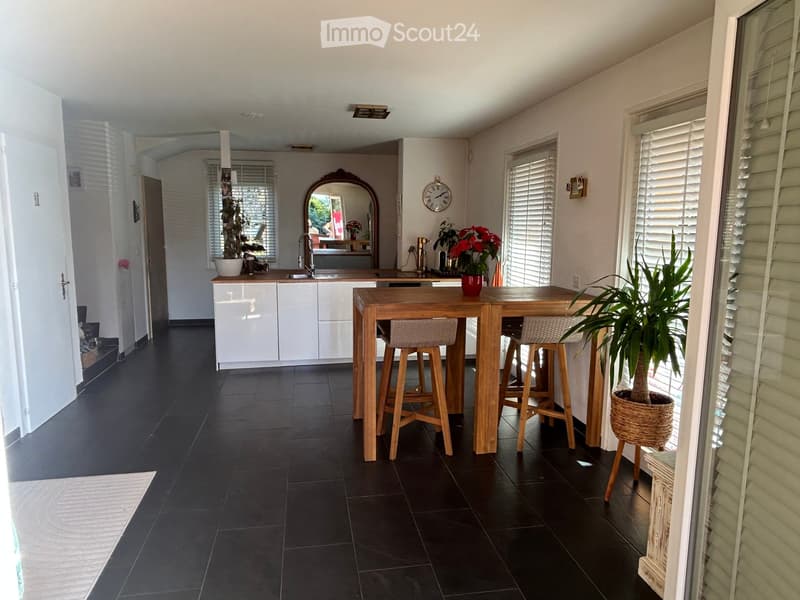 Modern kitchen with white cabinets, dark tiled floor, central island with bar stools, sink, and decorative plants.