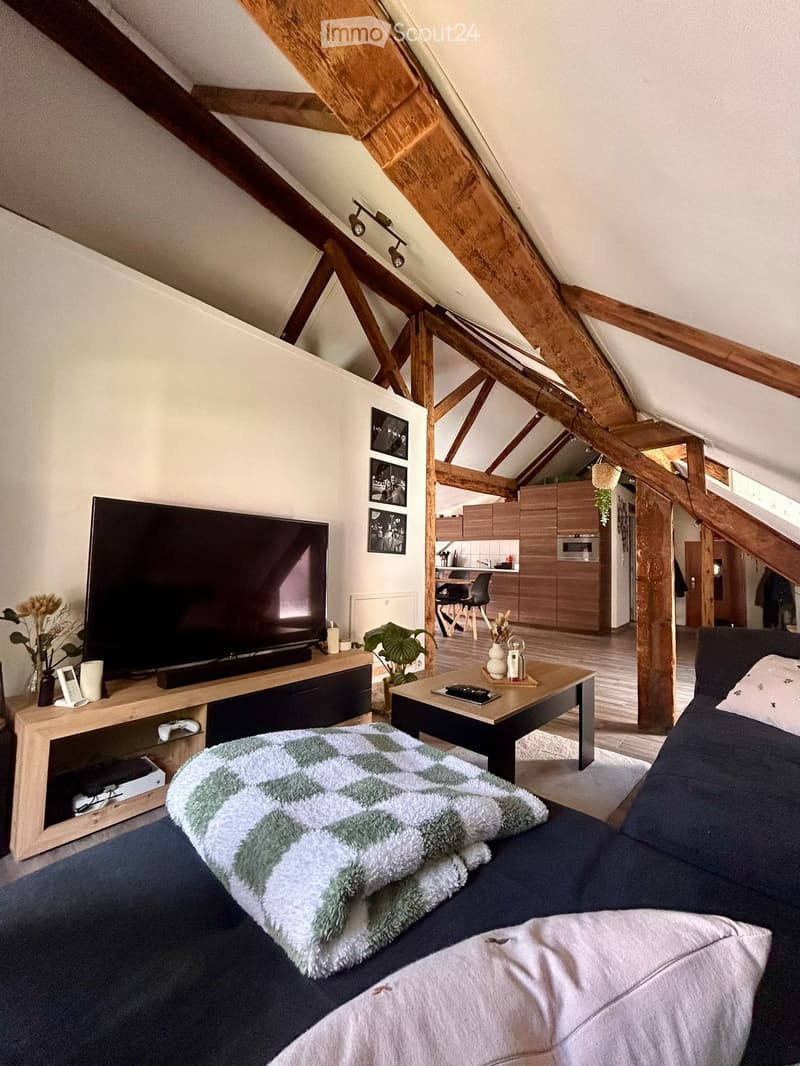 Cozy living room with exposed wooden beams, a sofa with a green and white blanket, a coffee table, a television on a wooden stand, and a small kitchen area with a microwave and chairs.