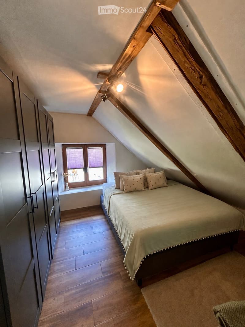 Cozy attic bedroom with wooden slanted ceilings, a large window, a neatly made bed with pillows, a wardrobe, and a small vase on the windowsill.